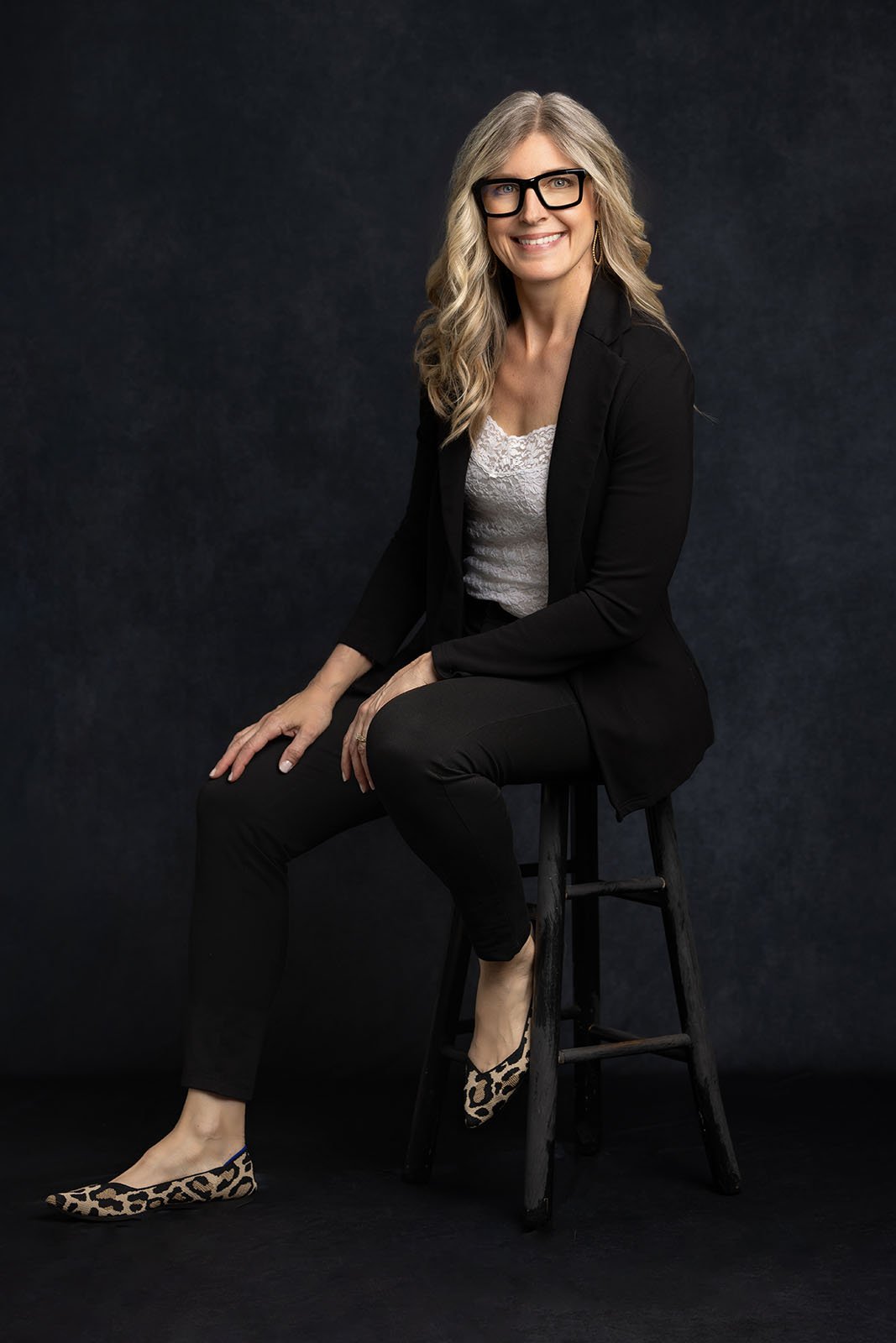 Woman in black sitting on stool in front of black studio backdrop wearing glasses and smiling for camera. This is in Julie Collins Photography's studio in Oconomowoc, Wisconsin